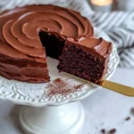 Moist chocolate cake with creamy frosting on a white floral cake stand, one slice lifted with golden cake server