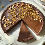 Overhead photo of a sliced raw vegan chocolate tart topped with nuts, cacao nibs, and bee pollen on a stone background in natural sunlight.