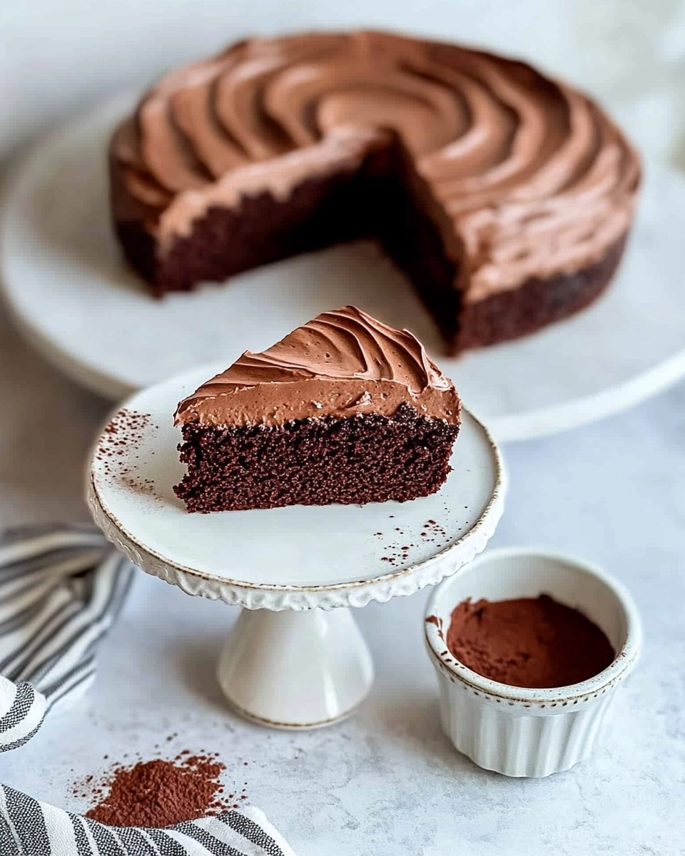 Chocolate cake slice with frosting in a ceramic bowl next to whole cake on a white stand