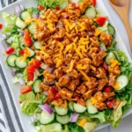Overhead view of a colorful BBQ chicken salad with fresh vegetables, cheese, and romaine lettuce on a white plate.