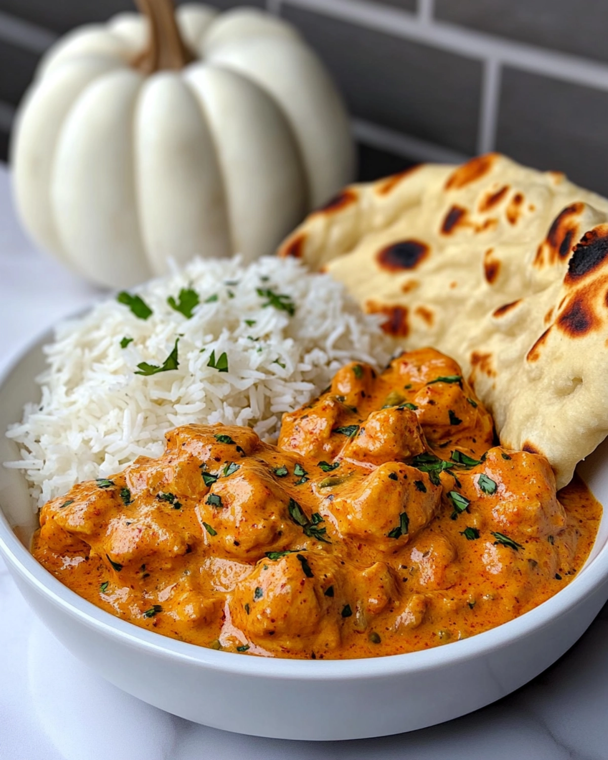 Bowl of creamy butter chicken with basmati rice and naan bread on a light background.