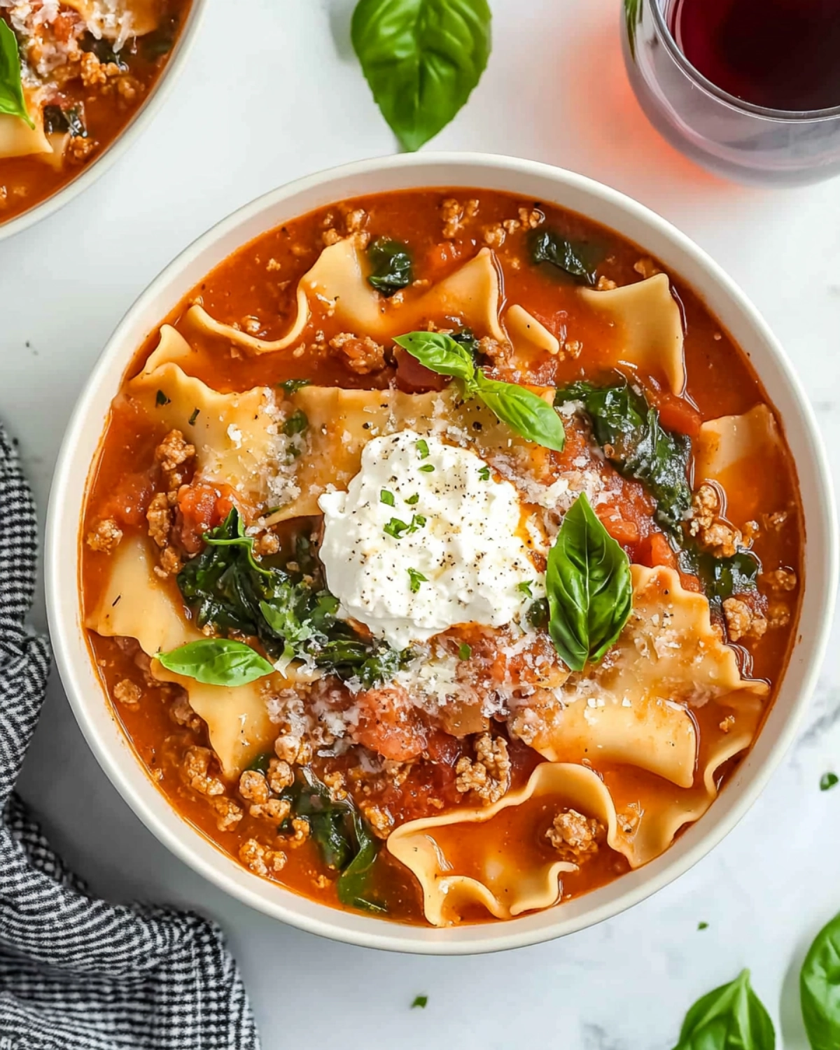 Bowl of lasagna soup with ricotta, basil, and parmesan, served on a marble background with red wine and a kitchen towel.