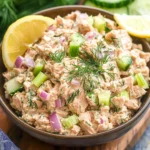 Close-up of creamy homemade tuna salad with red onion, celery, dill, and lemon wedges served in a bowl