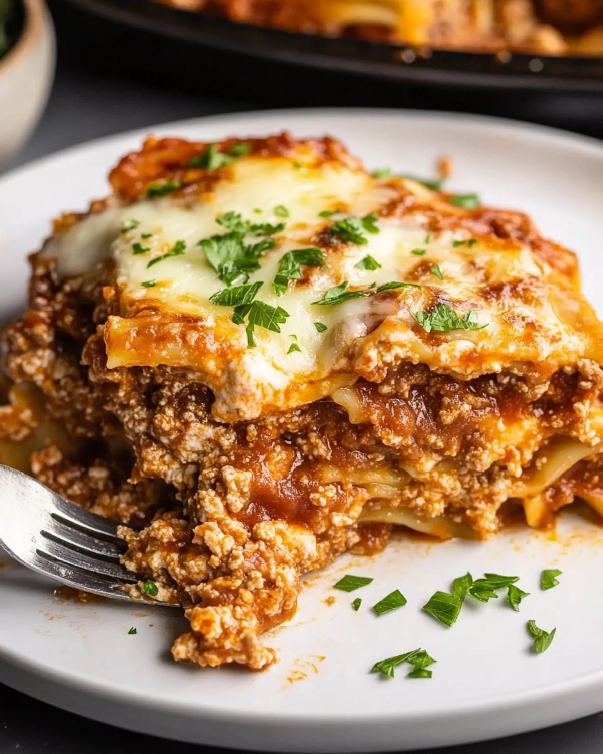 Close-up of a half-eaten lasagna slice on a white plate, showing layers of meat sauce, cheese, and pasta with a fork digging in.