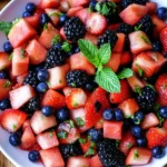 Colorful summer fruit salad with watermelon, strawberries, blueberries, and blackberries garnished with mint in a white bowl.