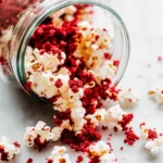 "Glass jar spilling red velvet popcorn onto a marble surface with crumbs scattered around."