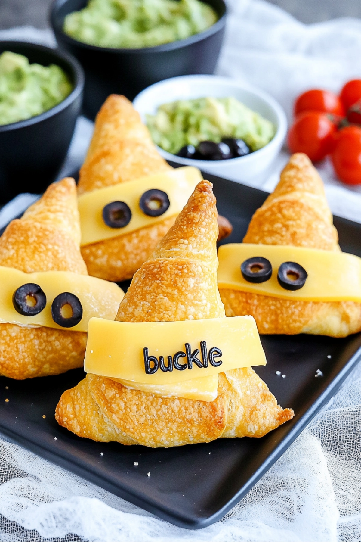 Hand holding a witch hat-shaped crescent roll with cheese and olive, served with guacamole, tomatoes, and olives.