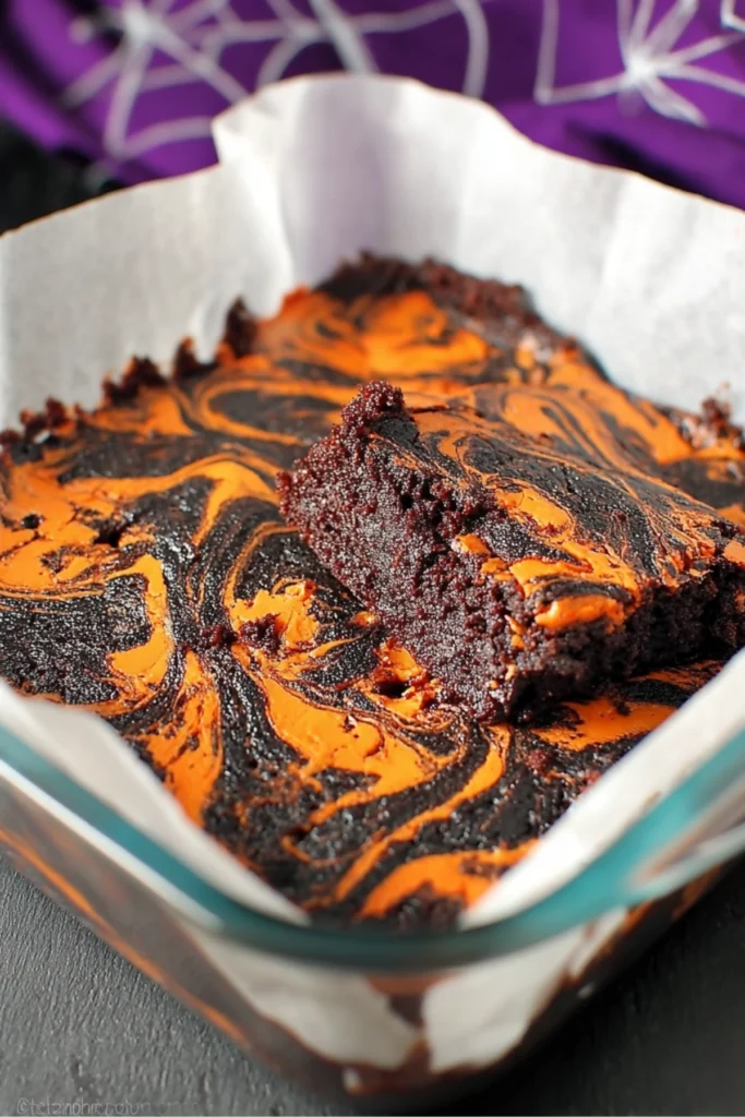 Halloween cheesecake brownies with black and orange swirls in a glass baking dish lined with parchment paper.