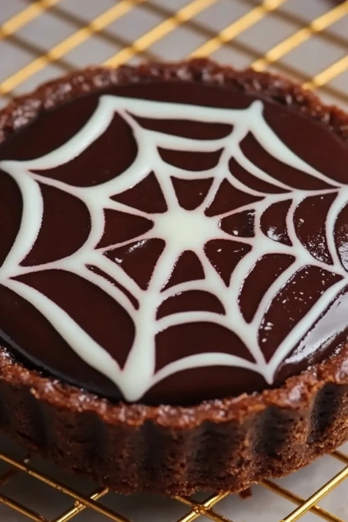 Chocolate tart with spiderweb icing design on ganache, placed on a golden cooling rack.