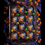 Halloween ghost brownies with candy toppings in a glass dish, surrounded by festive Halloween candy.