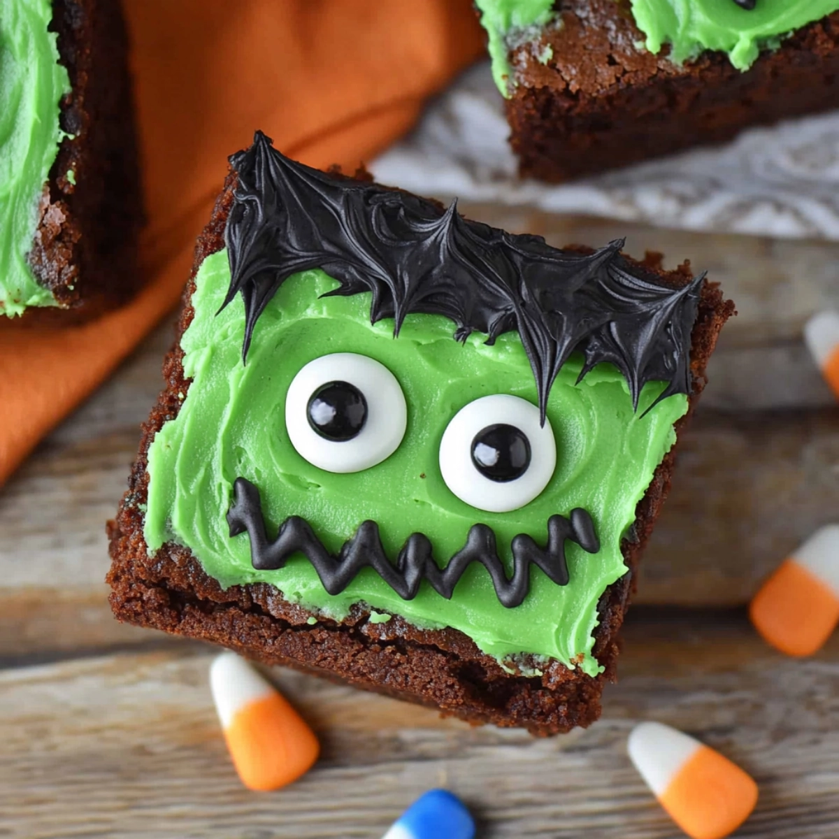 Halloween brownie decorated as Frankenstein's monster with green frosting, black icing hair, and candy eyes, surrounded by candy corn on a wooden surface.
