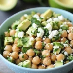 Fresh chickpea, avocado, and feta salad served in a blue bowl with lime and avocado in the background.