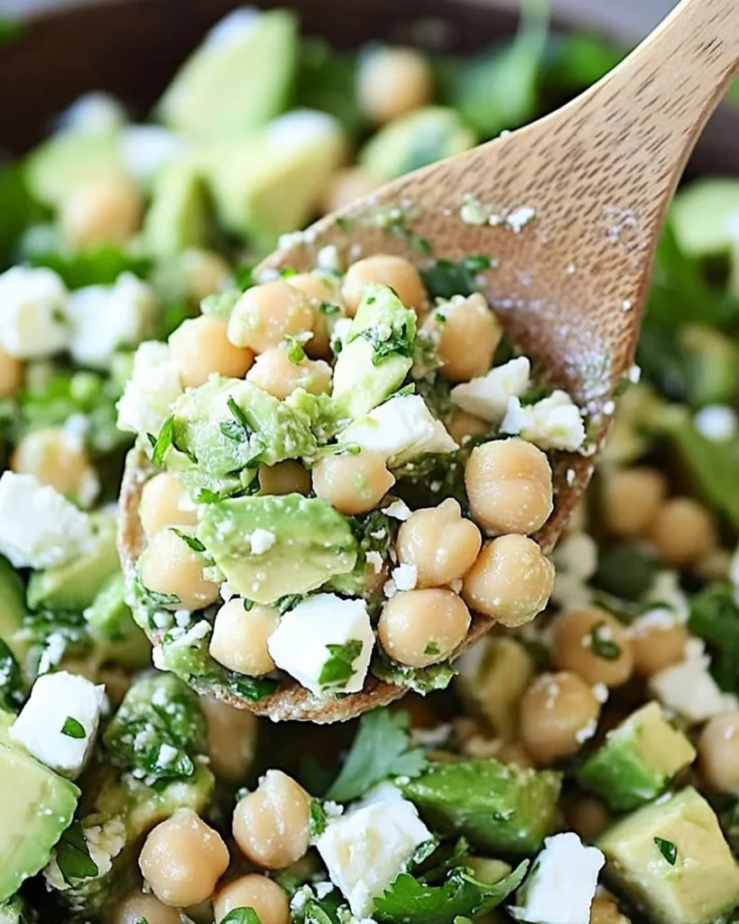 Close-up of chickpea, avocado, and feta salad on a wooden spoon, with fresh herbs and creamy textures.