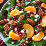 A fresh spinach salad with mandarin slices, pomegranate seeds, candied pecans, and feta cheese, beautifully arranged in a white bowl on a rustic wooden background.