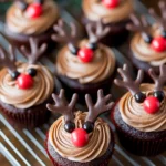 Chocolate reindeer cupcakes with candy eyes and antlers on a wire rack next to holiday decorations.