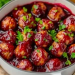 Cranberry glazed meatballs garnished with fresh parsley in a white pot, served in a rich red holiday sauce.