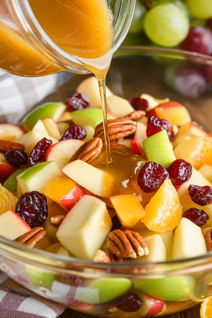 Hand pouring golden dressing over a vibrant fruit salad with apples, grapes, orange slices, cranberries, and pecans in a glass bowl.