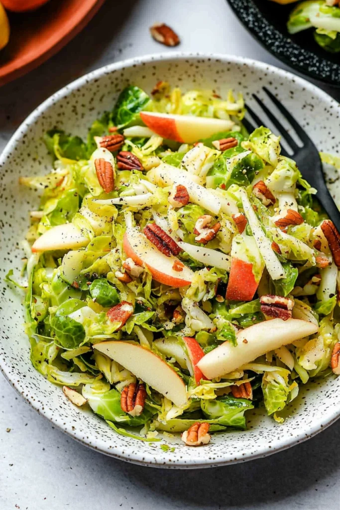 A fresh Brussels sprouts salad with apples, toasted nuts, and seeds in a speckled bowl, with a black fork placed on the side.