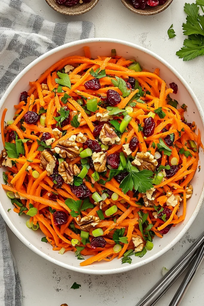 Colorful shredded carrot salad with walnuts, cranberries, green onions, and parsley served in a white bowl.