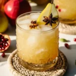 Festive fall cocktail with pear slice and star anise garnish, served in a textured glass on a woven coaster with pomegranates and pears in the background.
