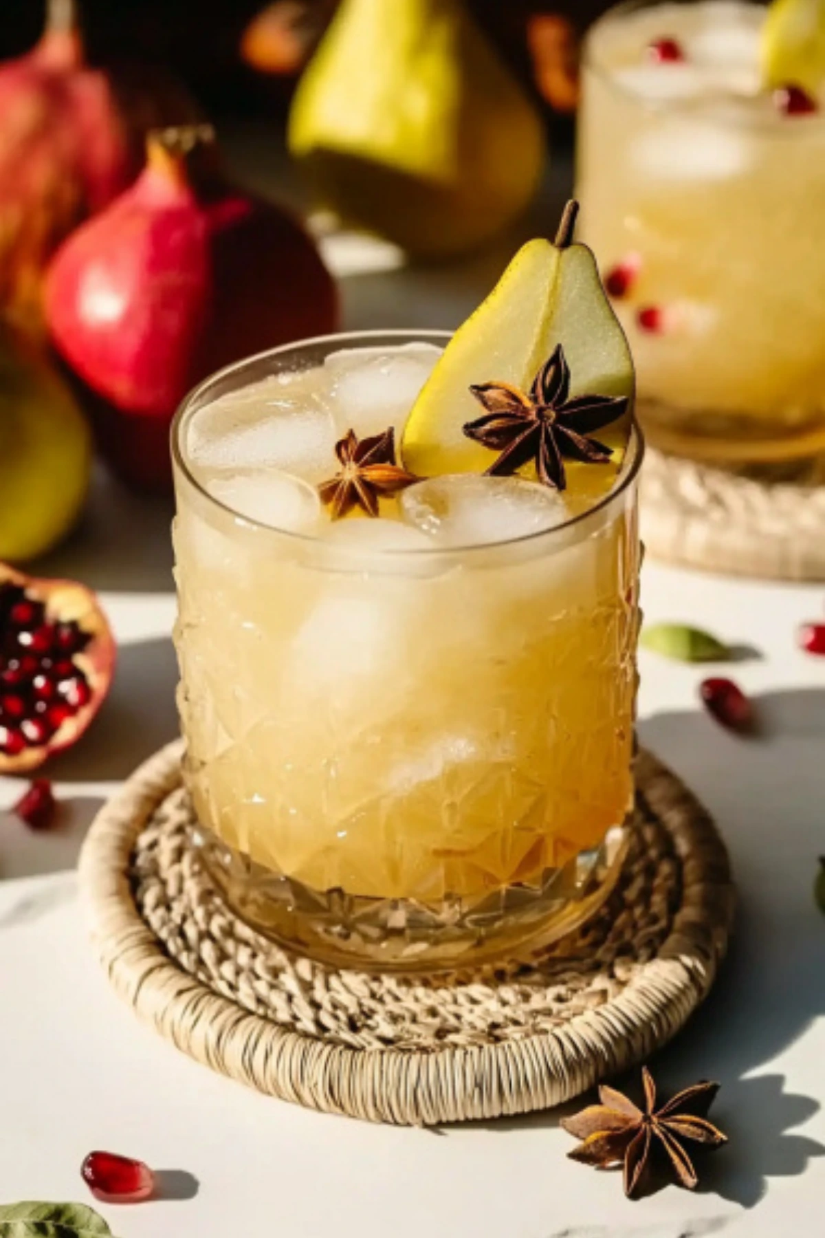 Festive fall cocktail with pear slice and star anise garnish, served in a textured glass on a woven coaster with pomegranates and pears in the background.