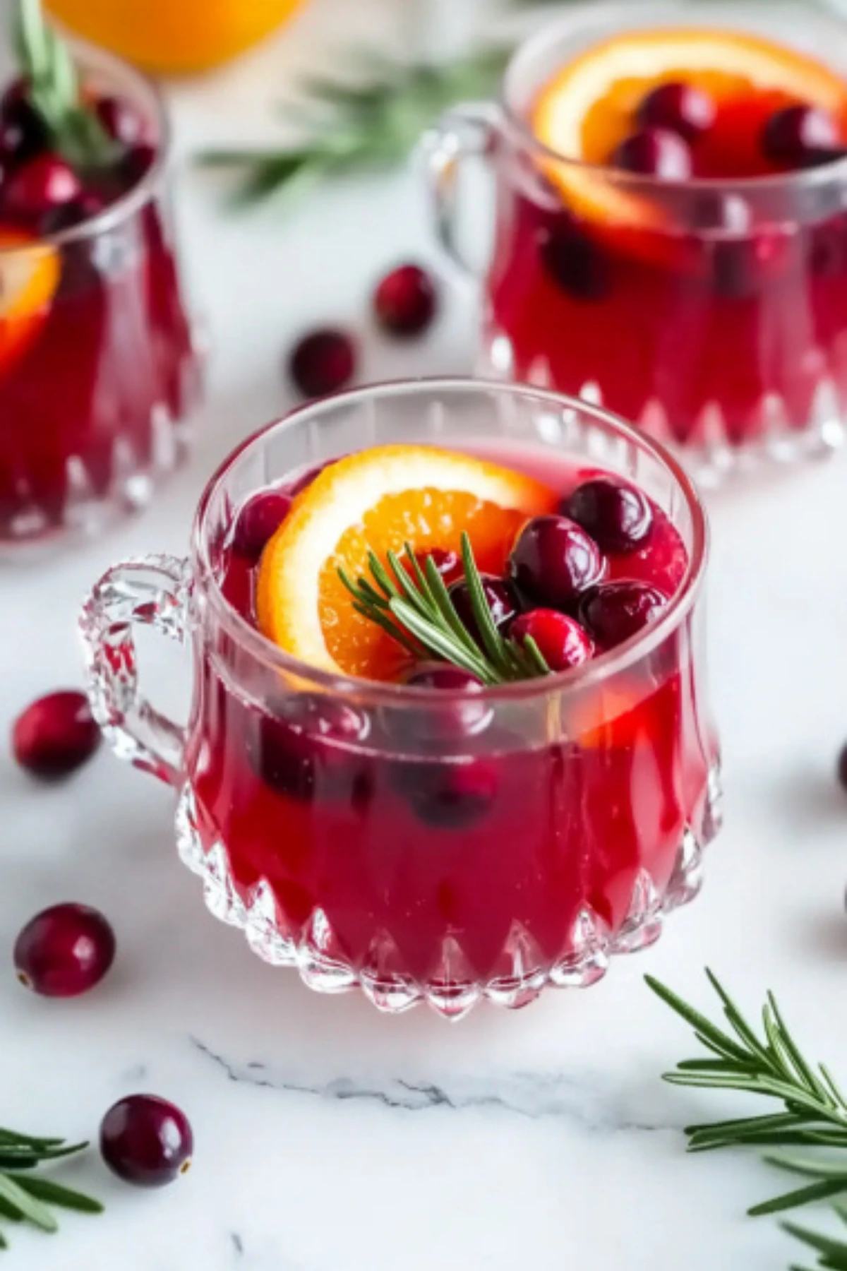 Clear glass cup filled with cranberry orange prosecco punch, garnished with fresh cranberries, orange slice, and rosemary sprig on white marble background