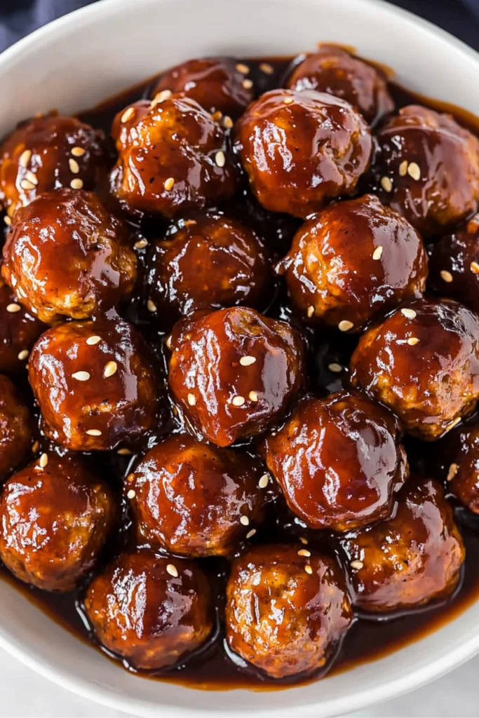 A close-up of glossy meatballs coated in a rich barbecue sauce, garnished with sesame seeds in a white bowl.