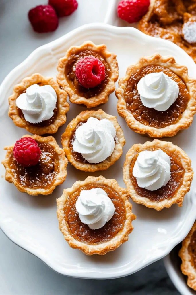 Mini pumpkin pies topped with whipped cream, with one pie bitten into to reveal the smooth pumpkin filling, set on a textured white plate.