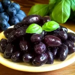 Close-up image of glossy dark purple grapes served on a white plate with basil leaves, with a bunch of grapes in the background.