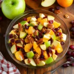 Healthy fruit salad with apples, grapes, orange slices, cranberries, and pecans in a clear bowl on a wooden surface.