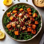 A vibrant salad with roasted sweet potatoes, kale, pomegranate, pumpkin seeds, and tahini dressing on a white background.