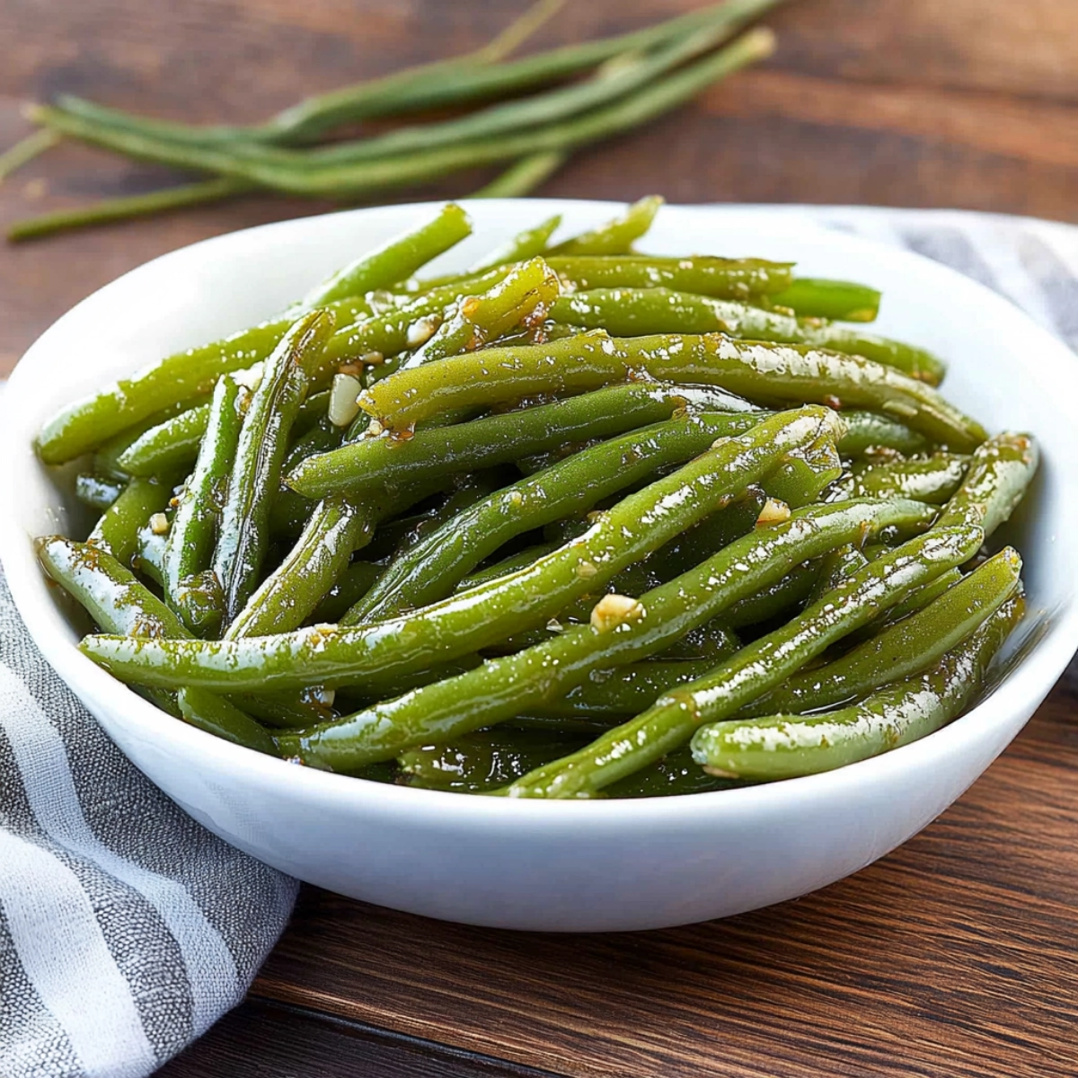 A close-up shot of sautéed green beans in a white bowl, with a hand holding the bowl, placed on a wooden surface, showcasing their glossy texture and vibrant green color.
