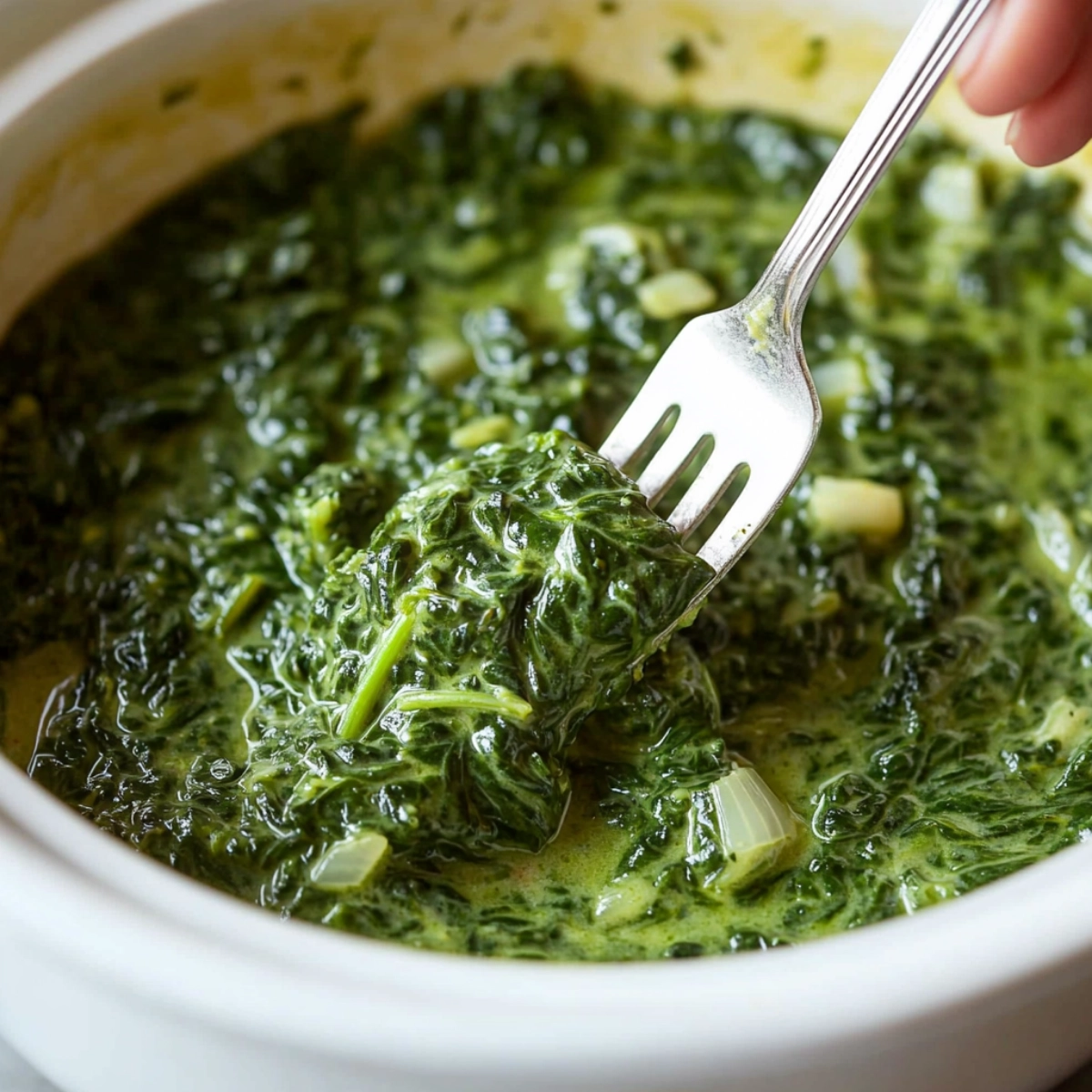 A close-up of creamy spinach being served with a fork from a crockpot, with a second shot of spinach topped with shredded cheese, featuring vibrant green color and smooth texture.