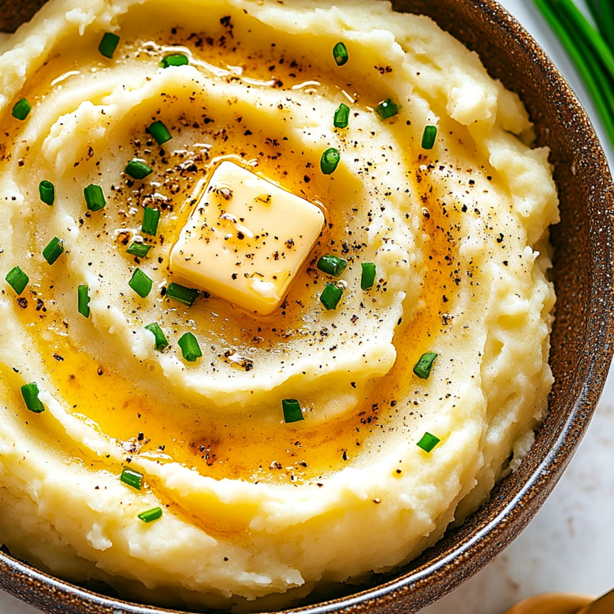 Closeup of creamy mashed potatoes with butter, garnished with chives and black pepper, served in a rustic ceramic bowl.