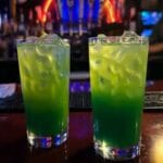 Green tropical cocktails with ice and vibrant layers in tall glasses, served at a bar with a neon light background.