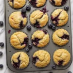 Freshly baked blueberry muffins in a muffin tin with blueberries on a marble counter.