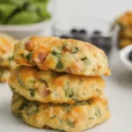 Stack of savory ham, cheese, and spinach biscuits on a white countertop with breakfast ingredients in the background.