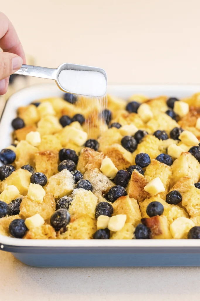 A hand pouring syrup over a slice of blueberry bread pudding on a white and blue plate with the full casserole dish in the background.