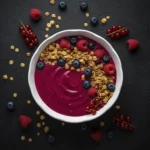 Vibrant pink berry smoothie bowl topped with blueberries, raspberries, mixed berries, and crunchy granola on a dark textured background.