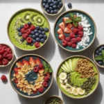 Close-up, overhead photograph of three vibrant green superfood smoothie bowls, generously topped with fresh berries (strawberries, raspberries, blueberries), sliced kiwi, toasted coconut flakes, and granola on a light background.
