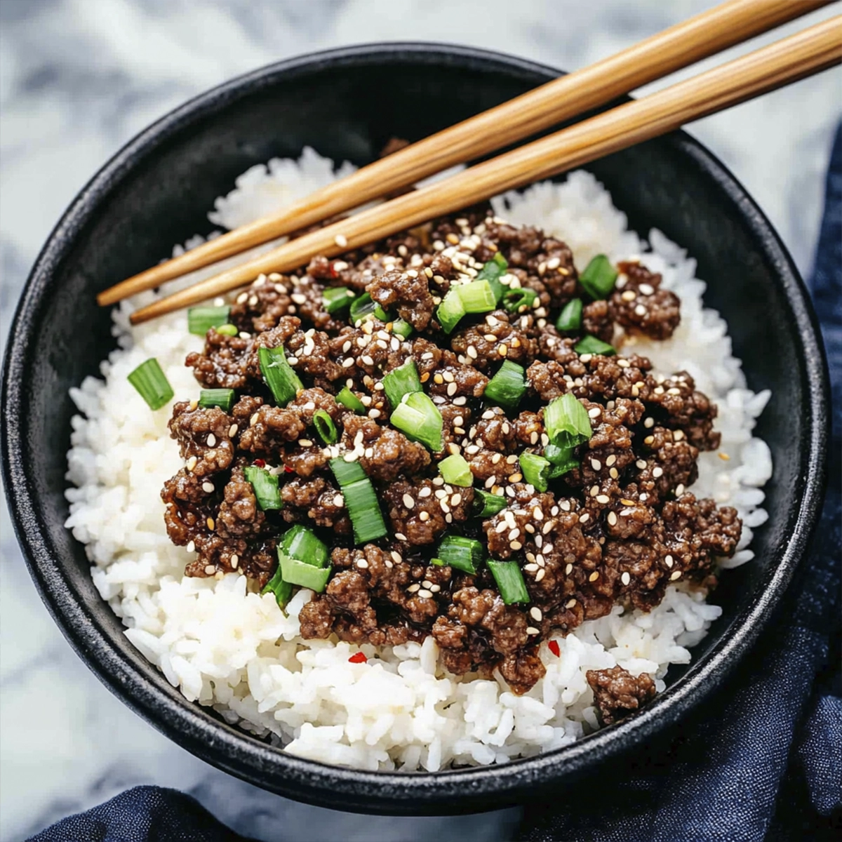 Korean-style ground beef with white rice topped with sesame seeds and green onions in a black bowl with chopsticks