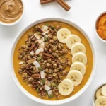 Overhead, bright flat lay photo of a pumpkin spice smoothie bowl in a white bowl, topped with granola, banana slices, and pumpkin seeds, surrounded by small dishes of toppings and cinnamon sticks.