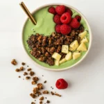 Green smoothie bowl with raspberries, granola, apples, chia seeds, and gold straw on white surface