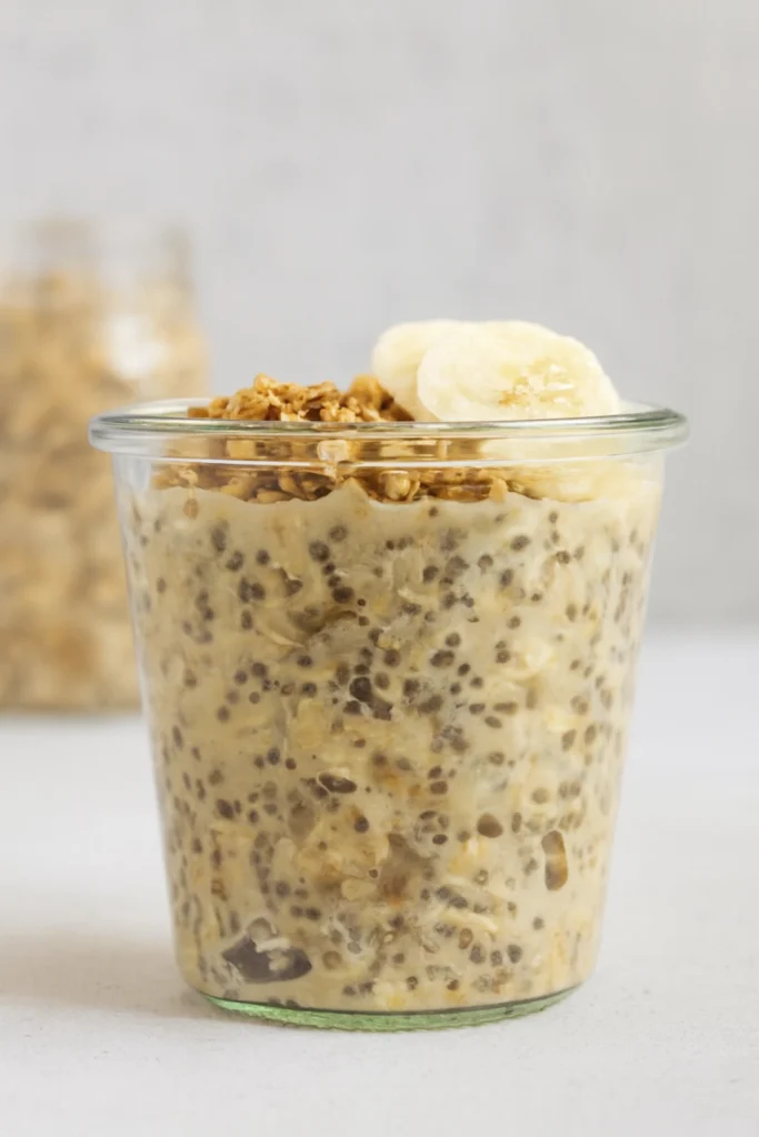Overnight oats with chia seeds, banana slices, and granola in a clear glass jar on a light background.
