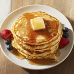 Fluffy stack of buttermilk pancakes topped with butter and maple syrup, served with fresh strawberries and blueberries.