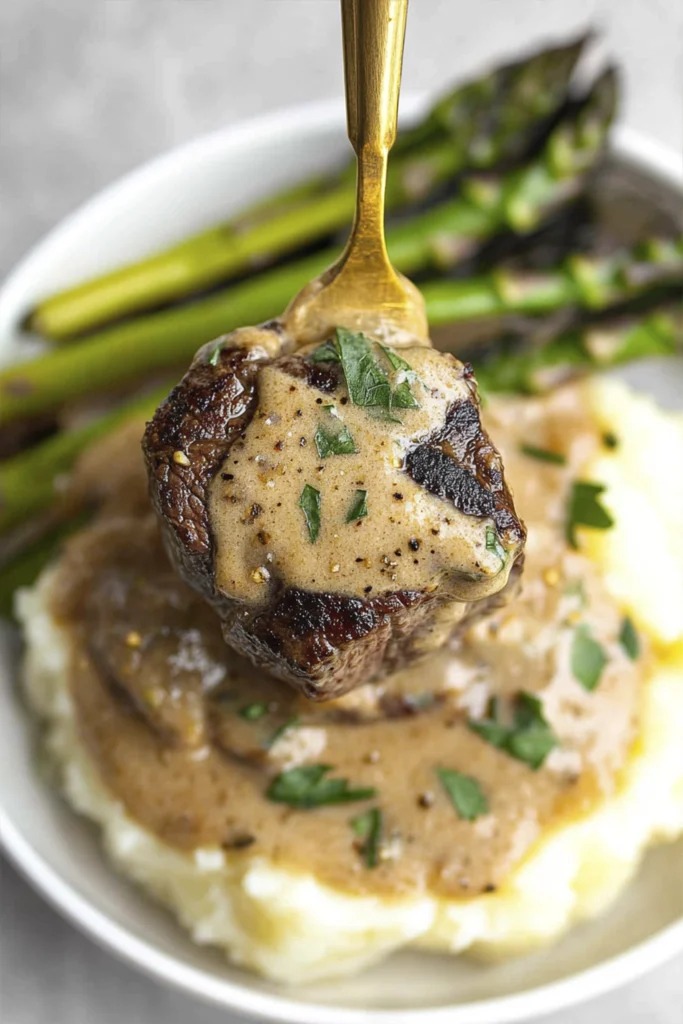 Close-up of creamy garlic steak bite lifted on a fork over mashed potatoes with asparagus on the side.