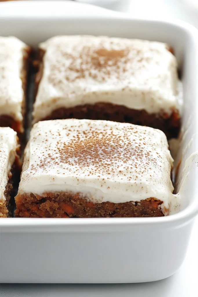 Carrot cake squares with cream cheese frosting in a white baking dish with one slice removed.