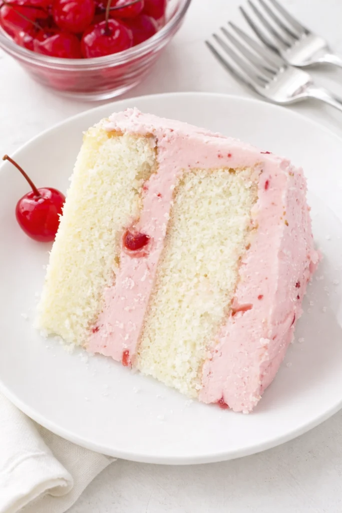 Slices of cherry layer cake with pink frosting and maraschino cherries served on white dessert plates