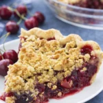 Close-up of a slice of cherry crumble pie with juicy cherry filling, buttery oat topping, and flaky crust on a white plate.