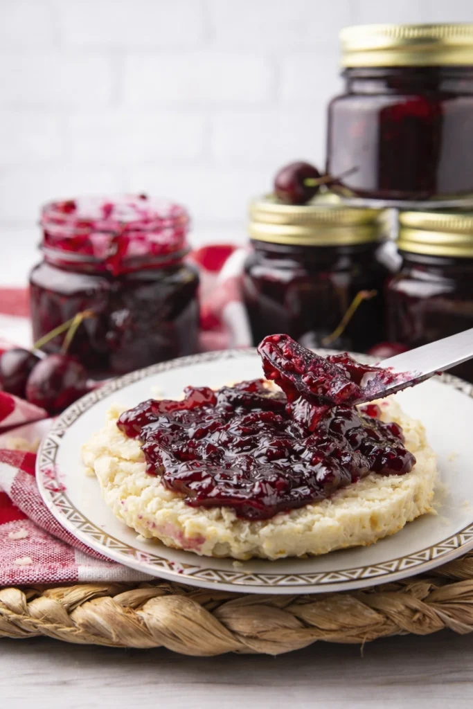 Homemade cherry jam spread on a biscuit with jars of cherry preserves and fresh cherries in the background